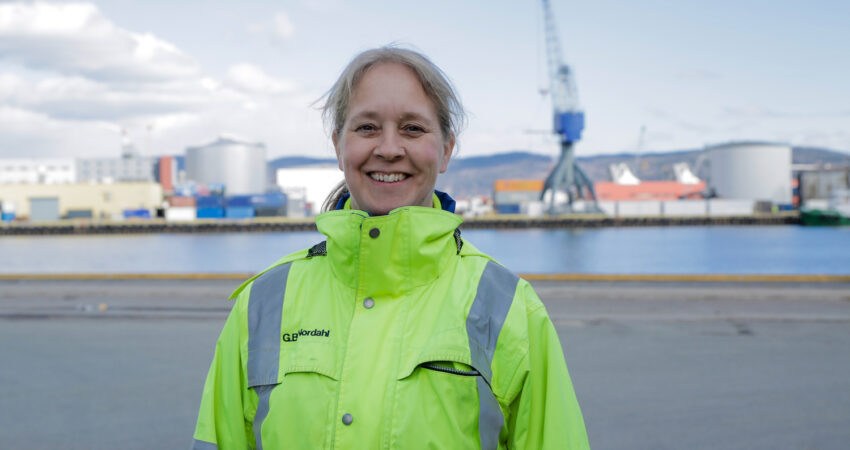 Gudveig nordahl staar paa kaia i gul arbeidsjakke. Vannet og kran i bakgrunnen.